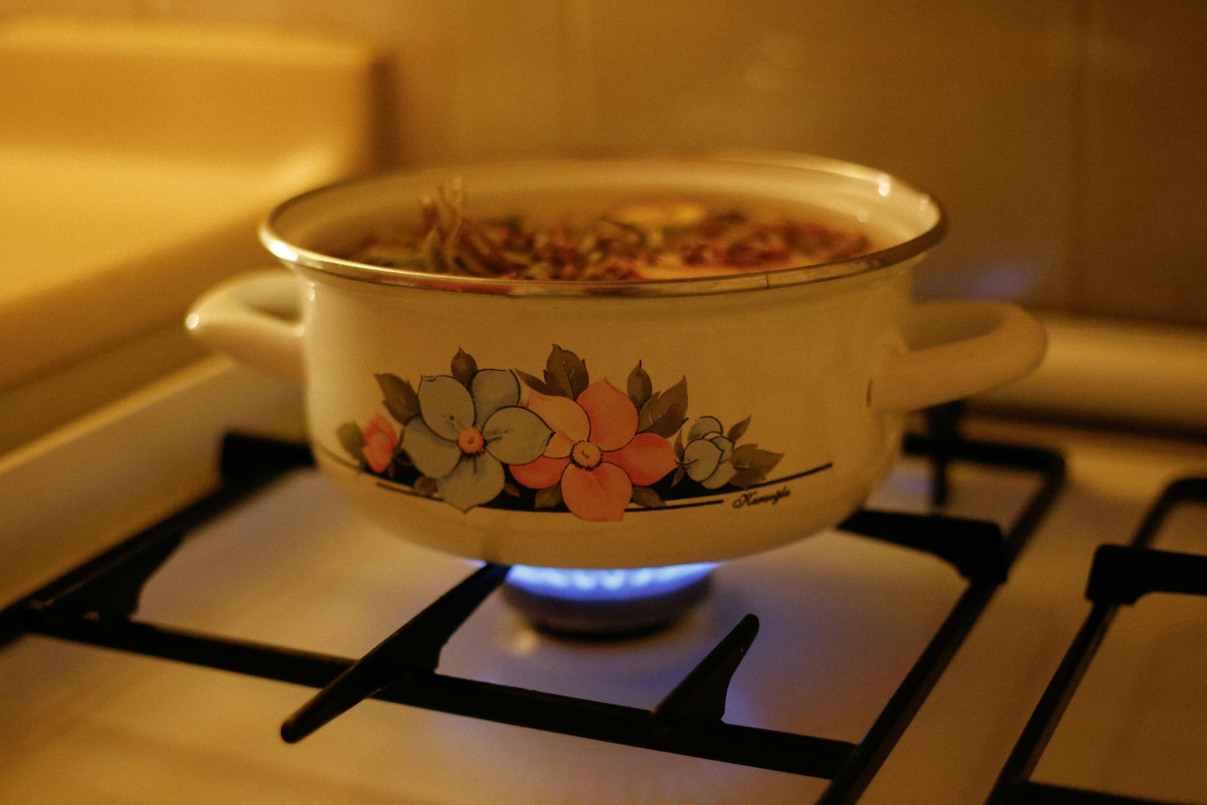 A close-up of a white ceramic pot with colorful floral designs, sitting on a gas stove with a blue flame underneath.